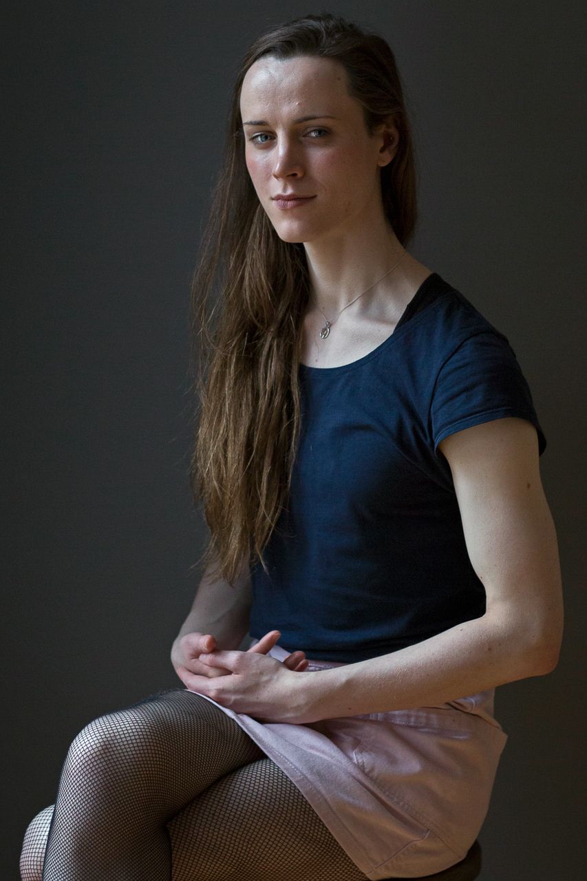 Young woman with long dark hair, wearing a dark shirt, sits looking at camera.