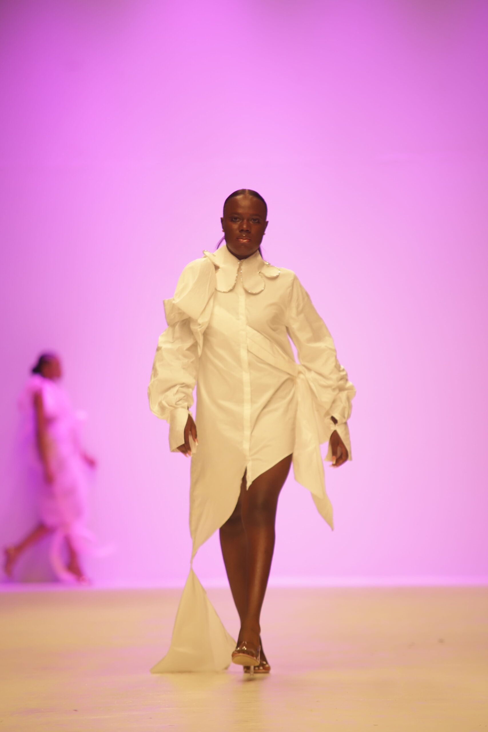 Dark-skinned model wears an avant-garde white shirt dress on a purple runway.