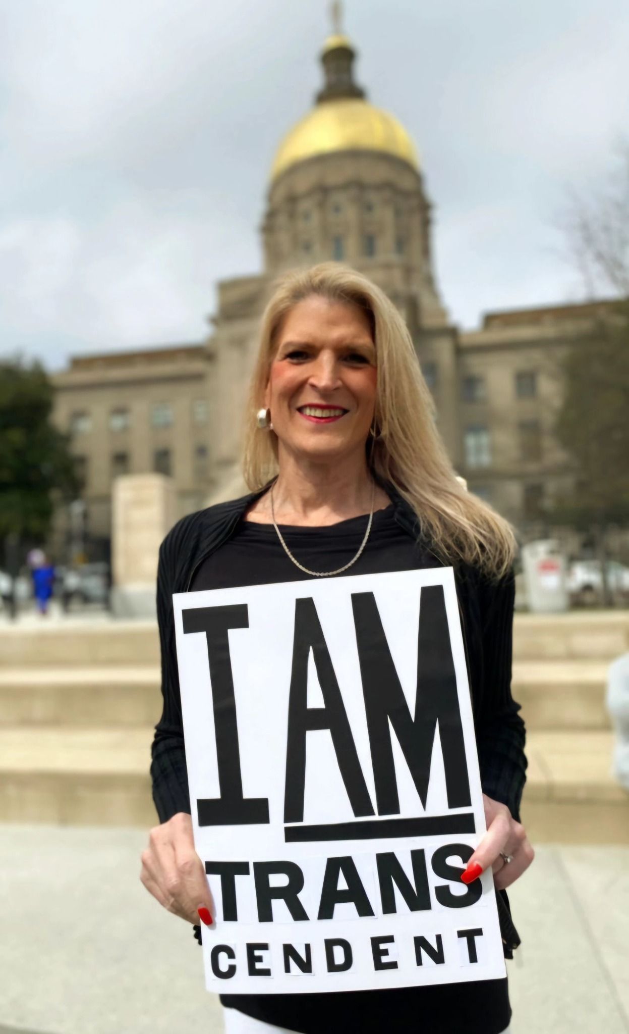 Person holds 'I AM TRANSCENDENT' sign on steps of a state capitol building.
