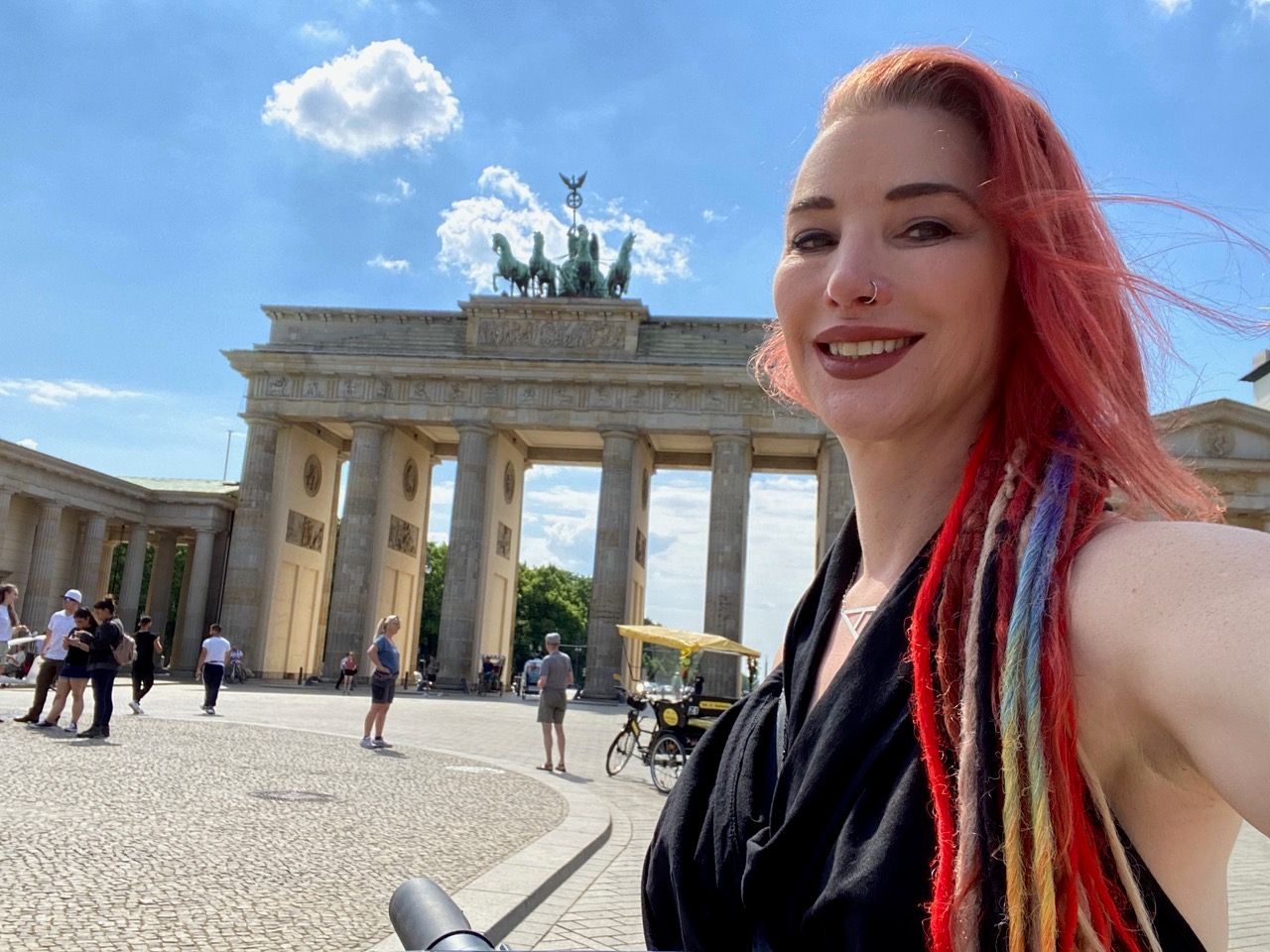 Smiling woman with red hair and dreadlocks taking a selfie in front of Brandenburg Gate, Berlin.