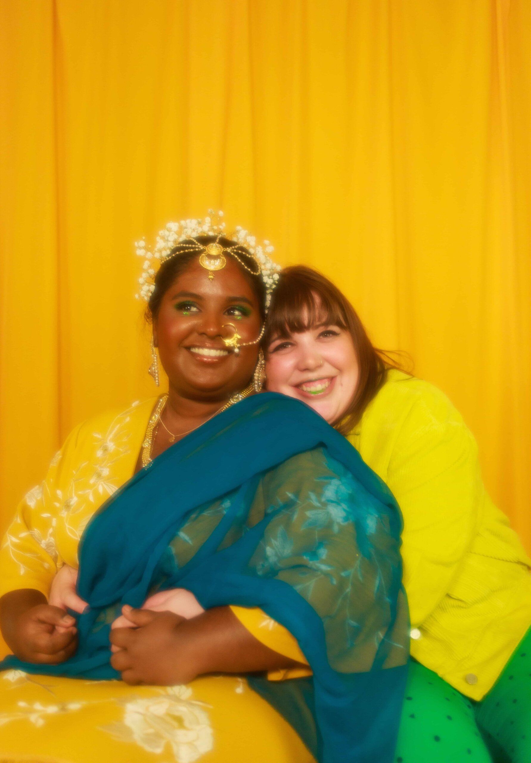 Two smiling women, one in traditional attire, embrace against a vibrant yellow background.