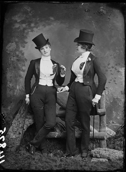 Two women in tuxedos and top hats stand posed in a vintage studio photograph.