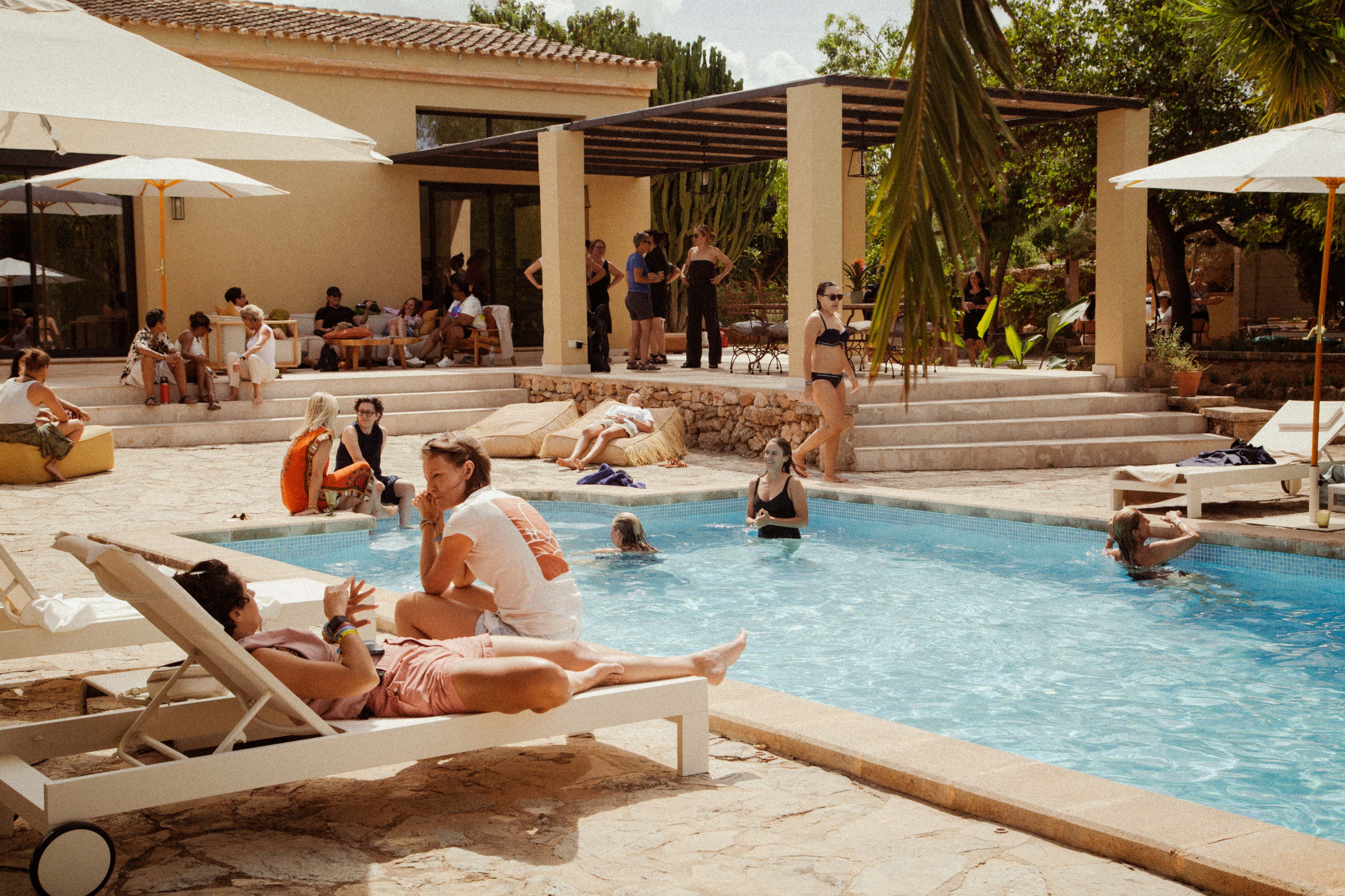 People relaxing by a large swimming pool at a sunny villa with palm trees.