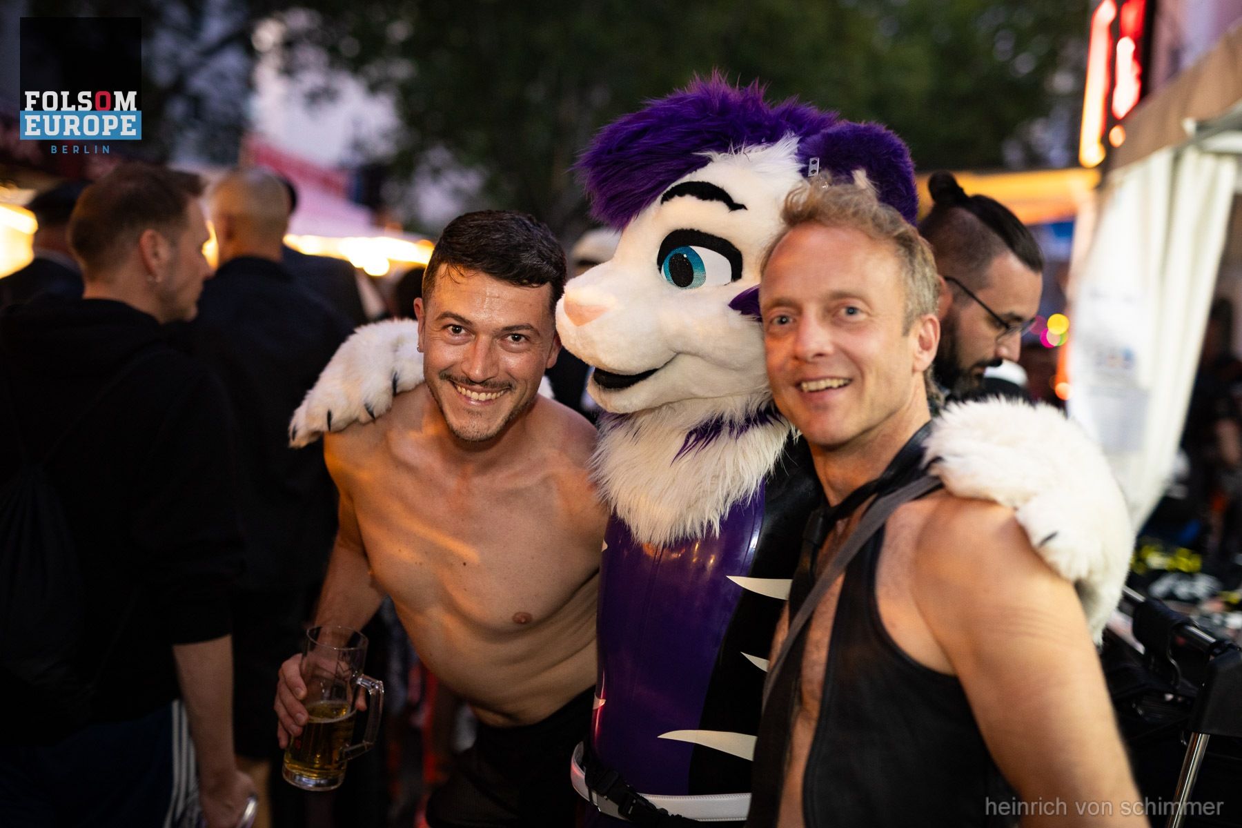 Two smiling men and a person in a purple furry costume pose at a Folsom Europe event.