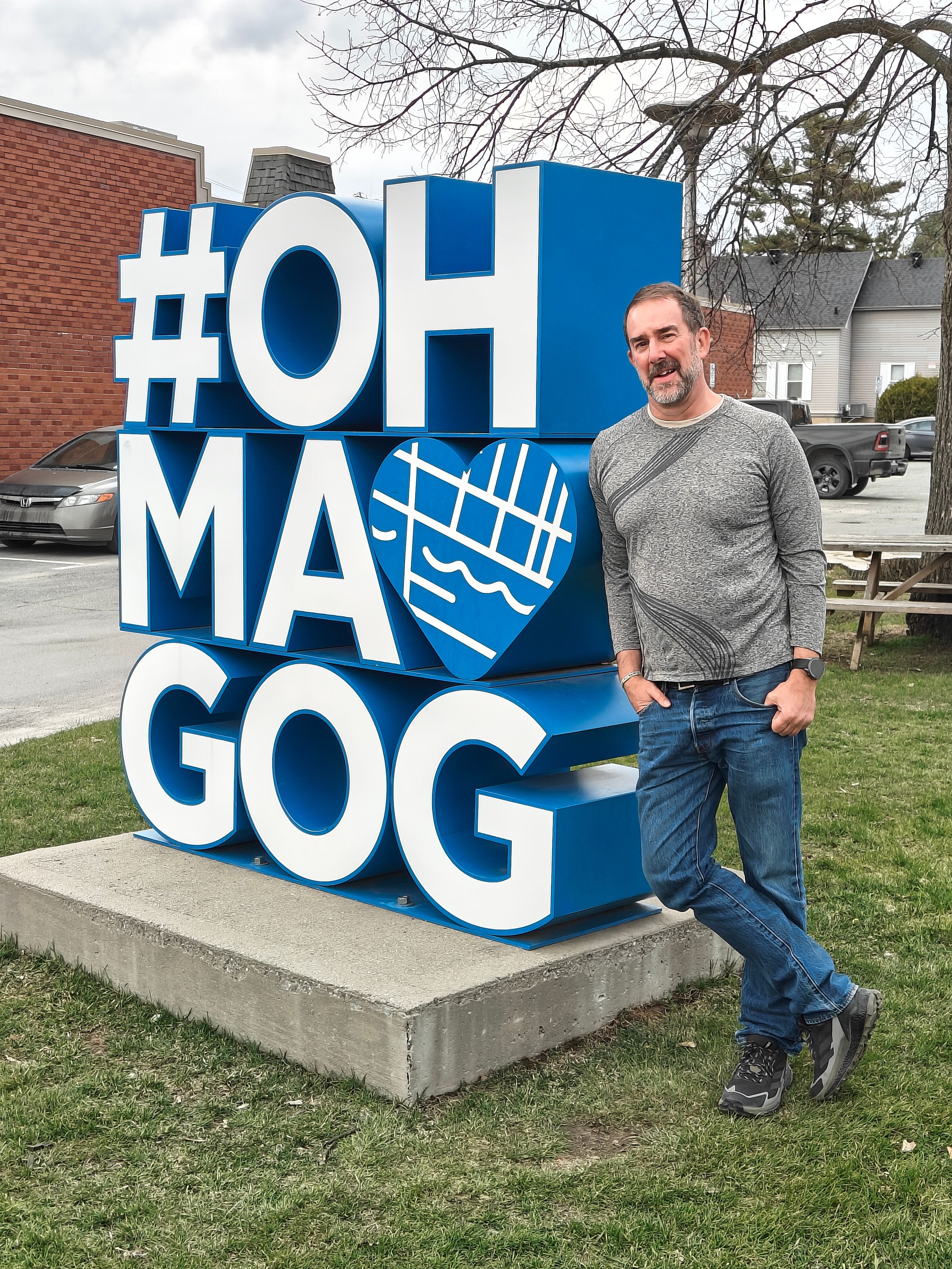 Man leans on a large blue and white '#OH MAGOG' sculpture featuring a heart map.