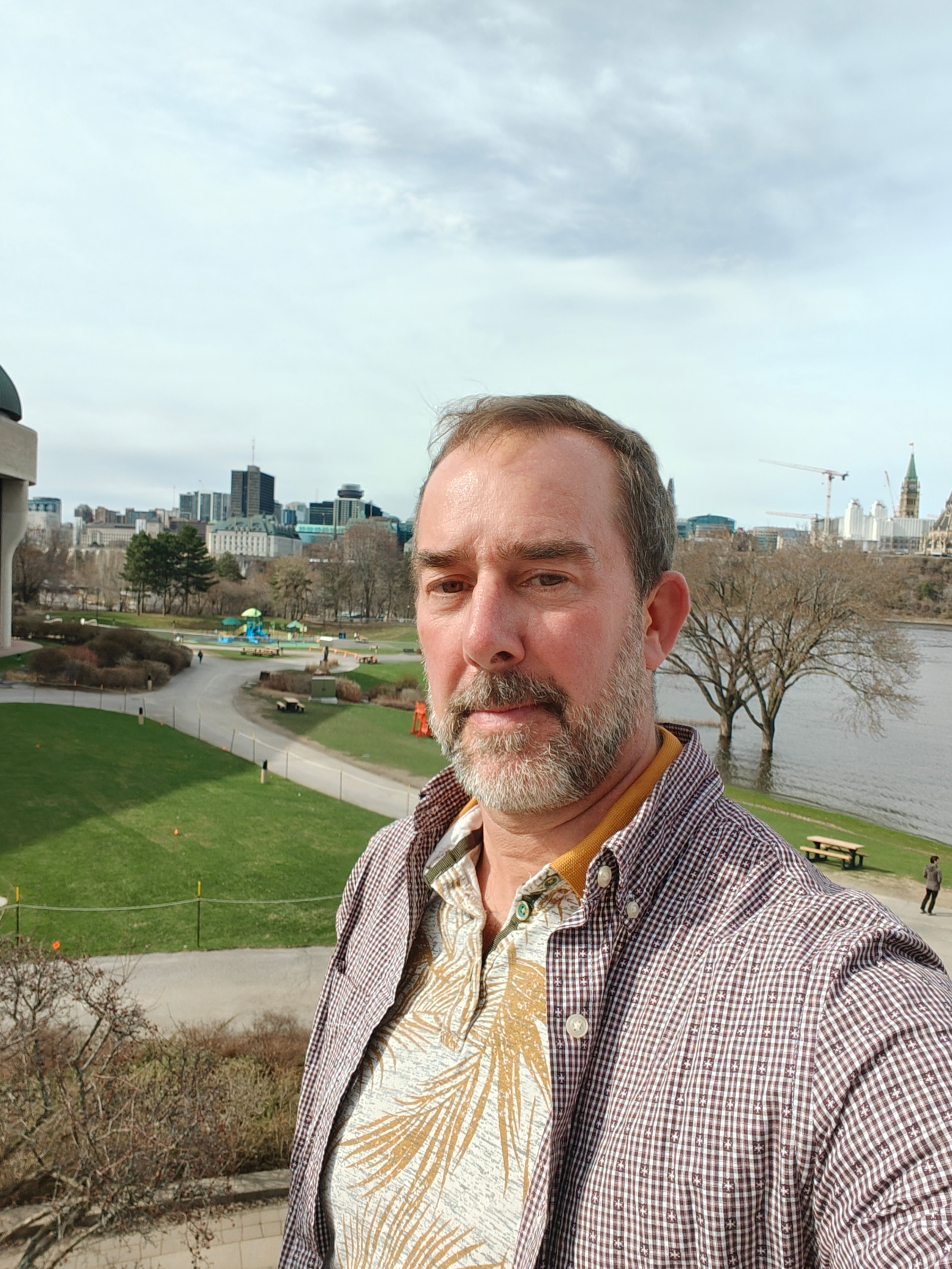 A bearded man takes a selfie outdoors with a city, river, and park behind him.