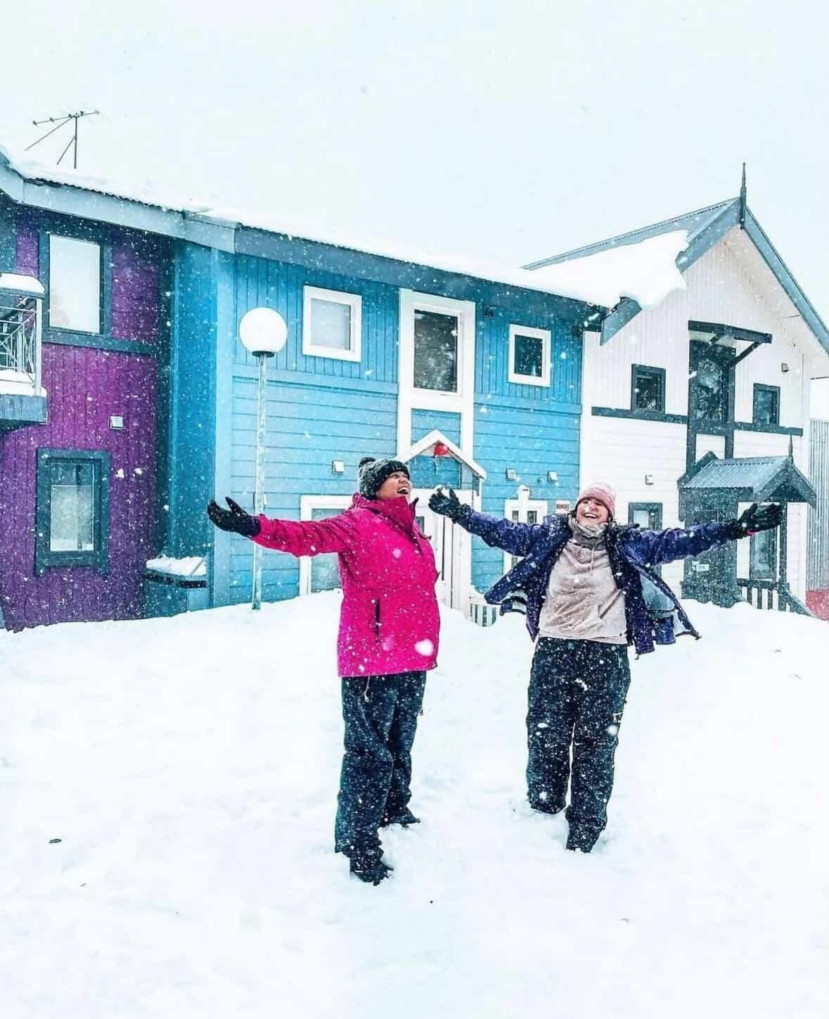 Two joyful people play in fresh snow, arms outstretched, before colorful houses.