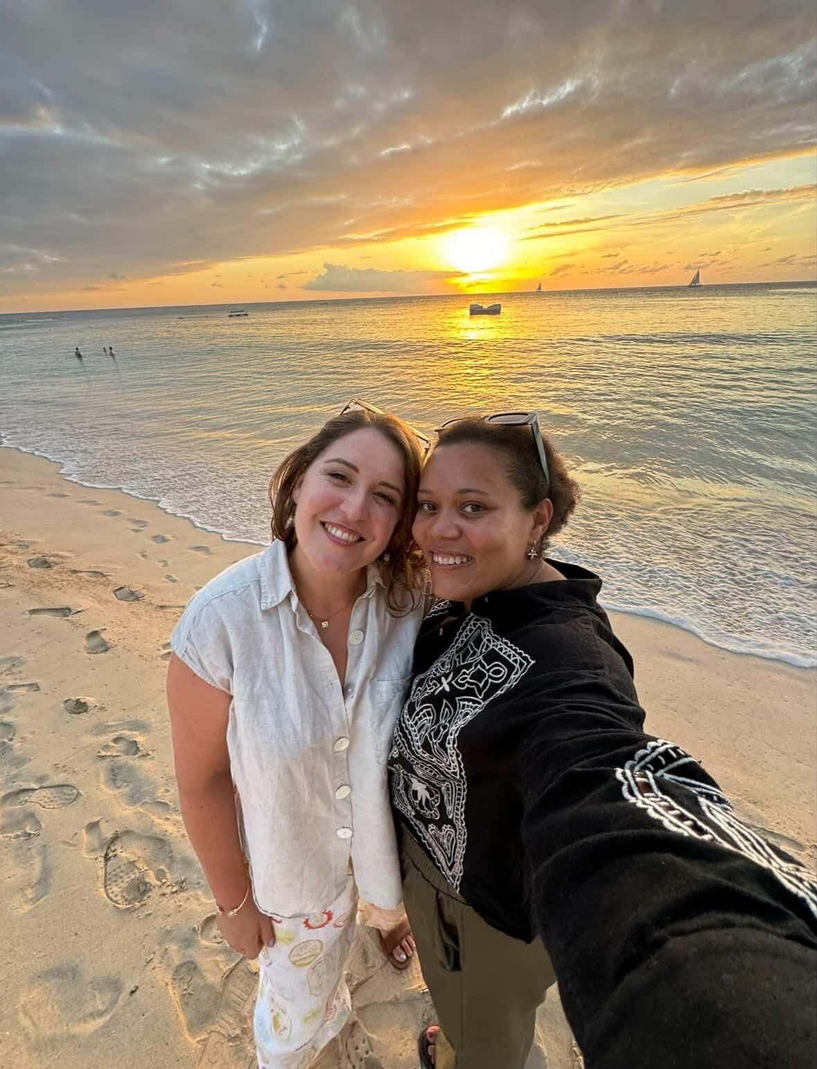 Two smiling women pose on a beach at sunset with a vibrant sky and ocean.