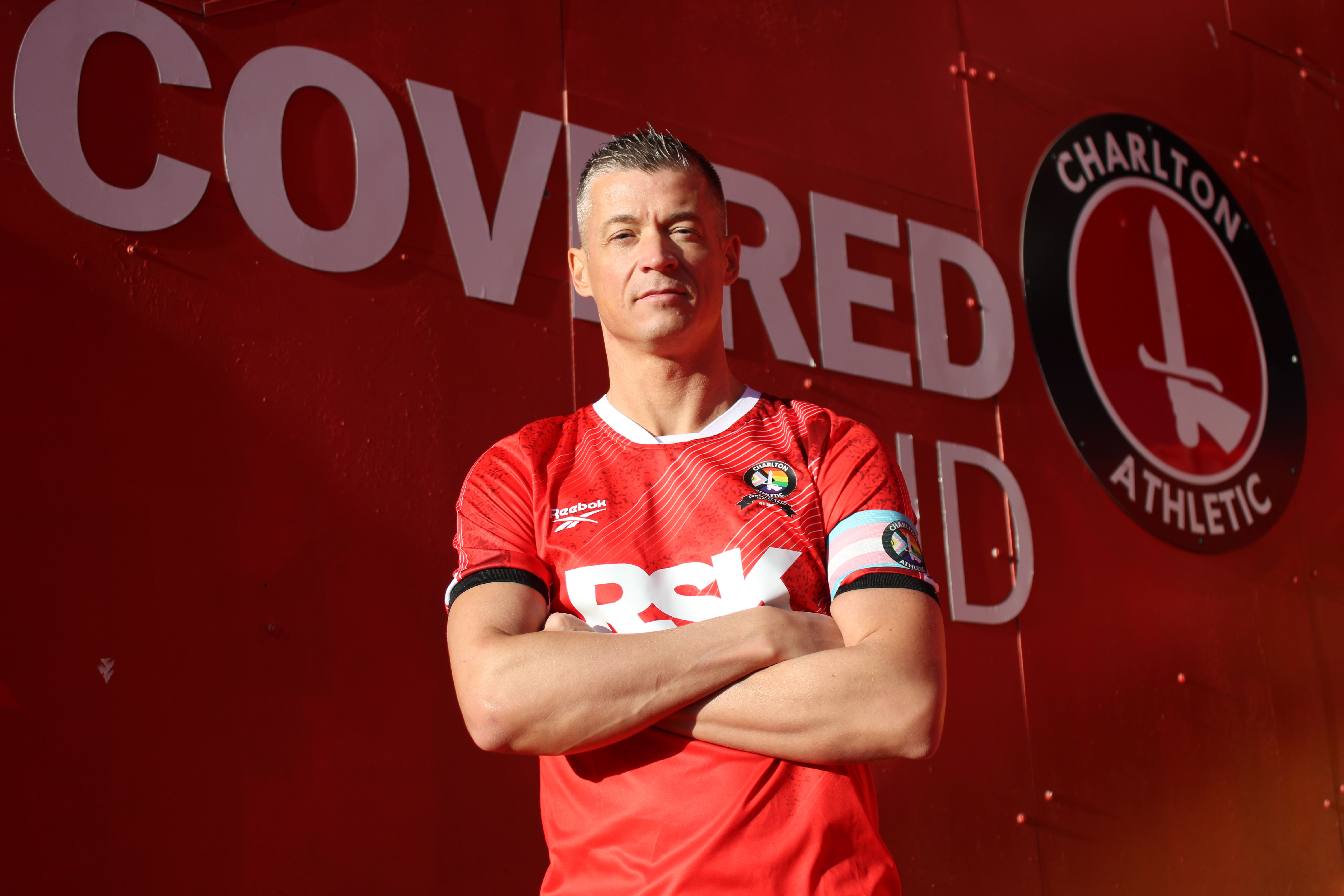 Gary Ginnaw in red Charlton Athletic jersey, arms crossed, standing confidently against a red banner.