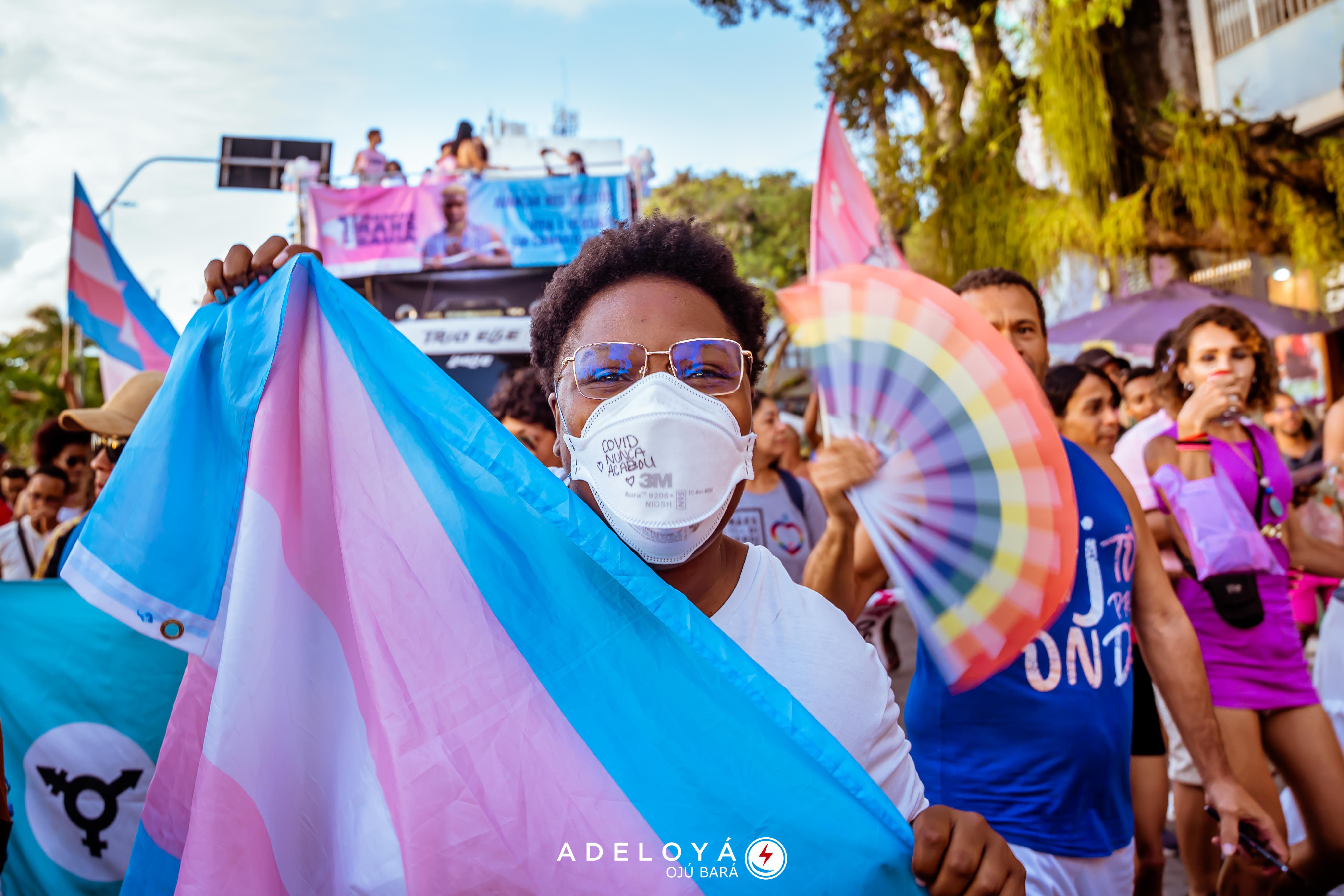 Masked person in glasses holds a transgender pride flag at a vibrant outdoor rally.