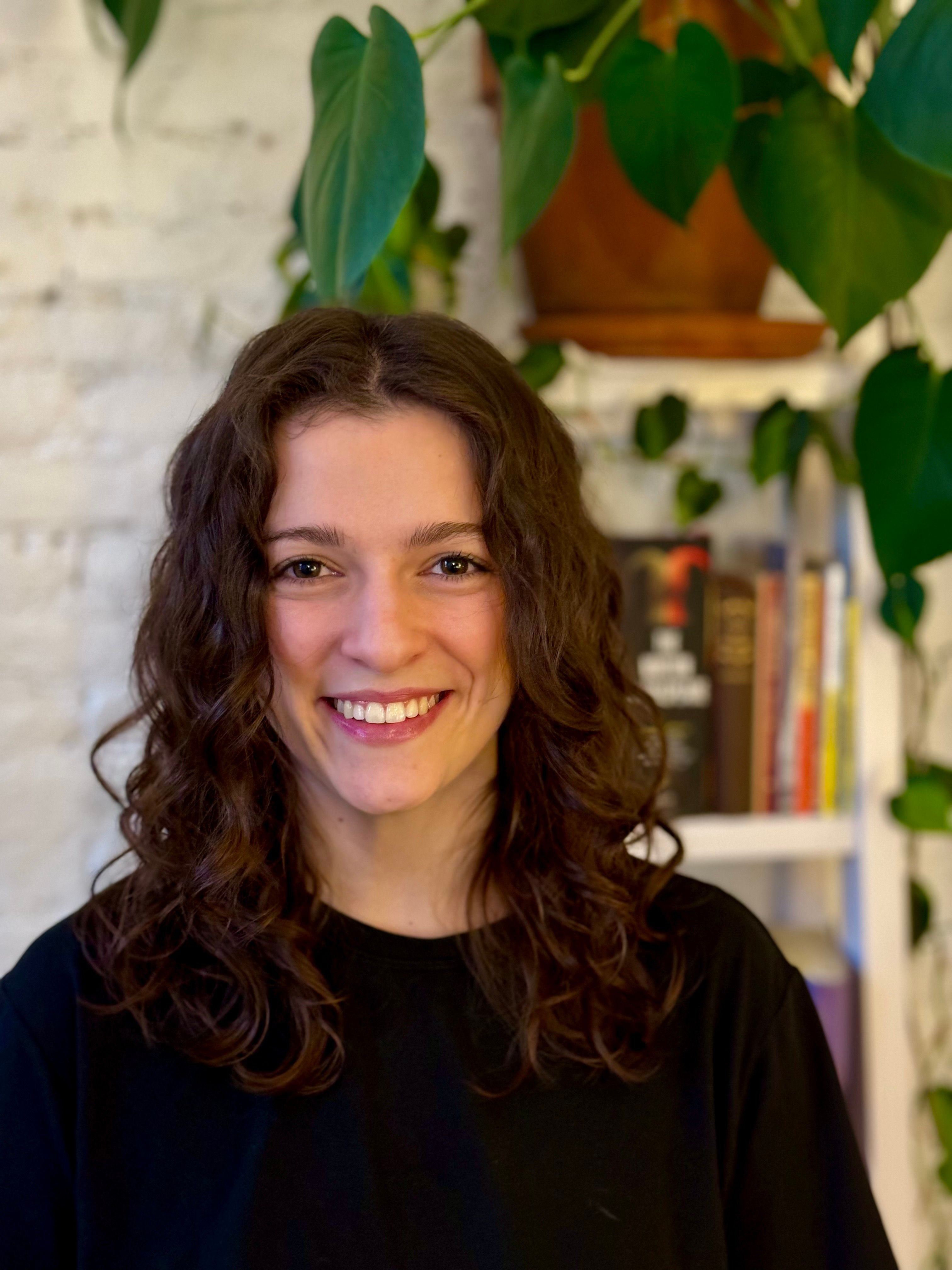 Smiling young woman with curly dark hair stands against a green plant background.