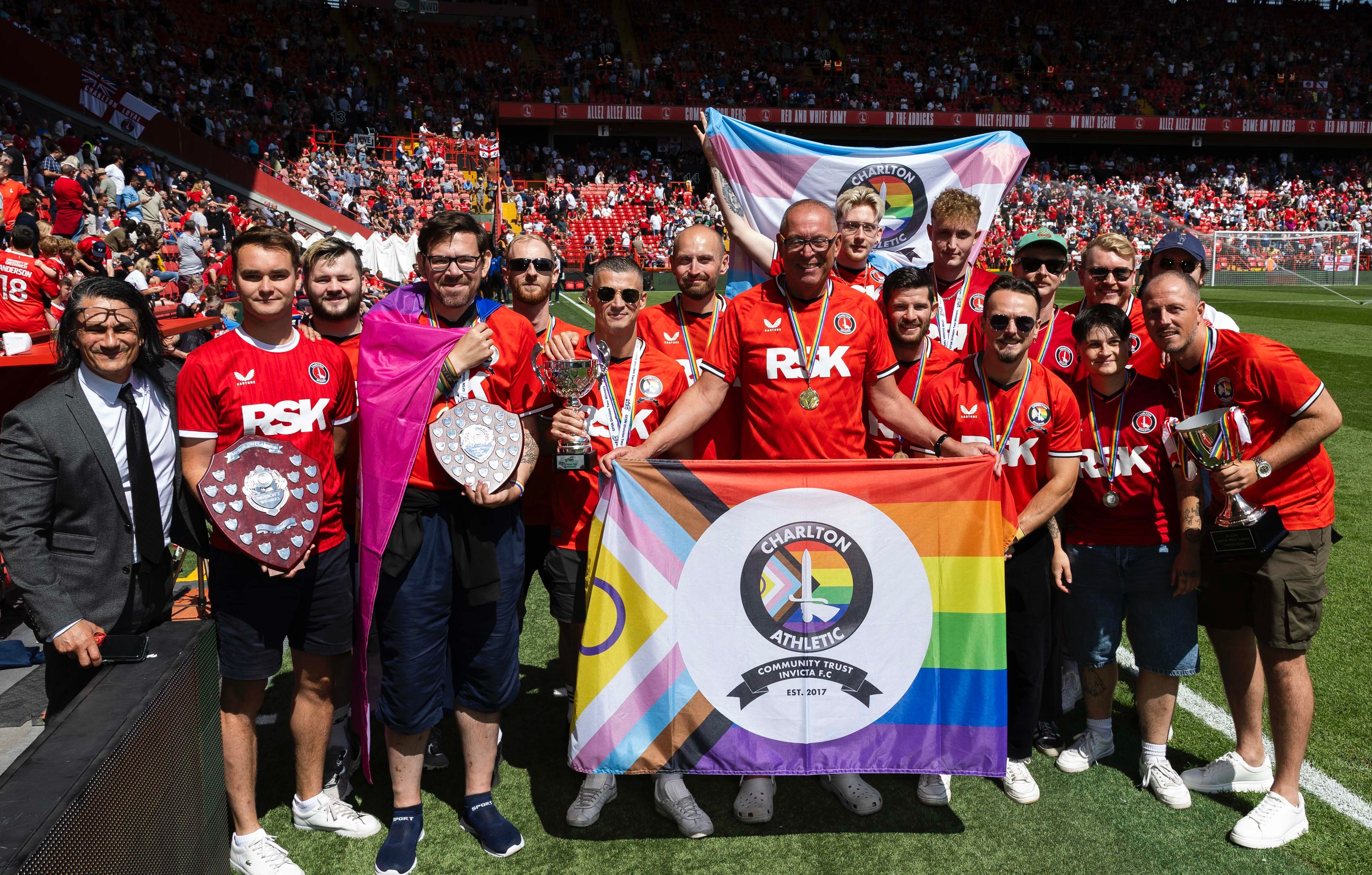 Gary Ginnaw Men in red jerseys hold trophies and various pride flags on a sunny soccer pitch.