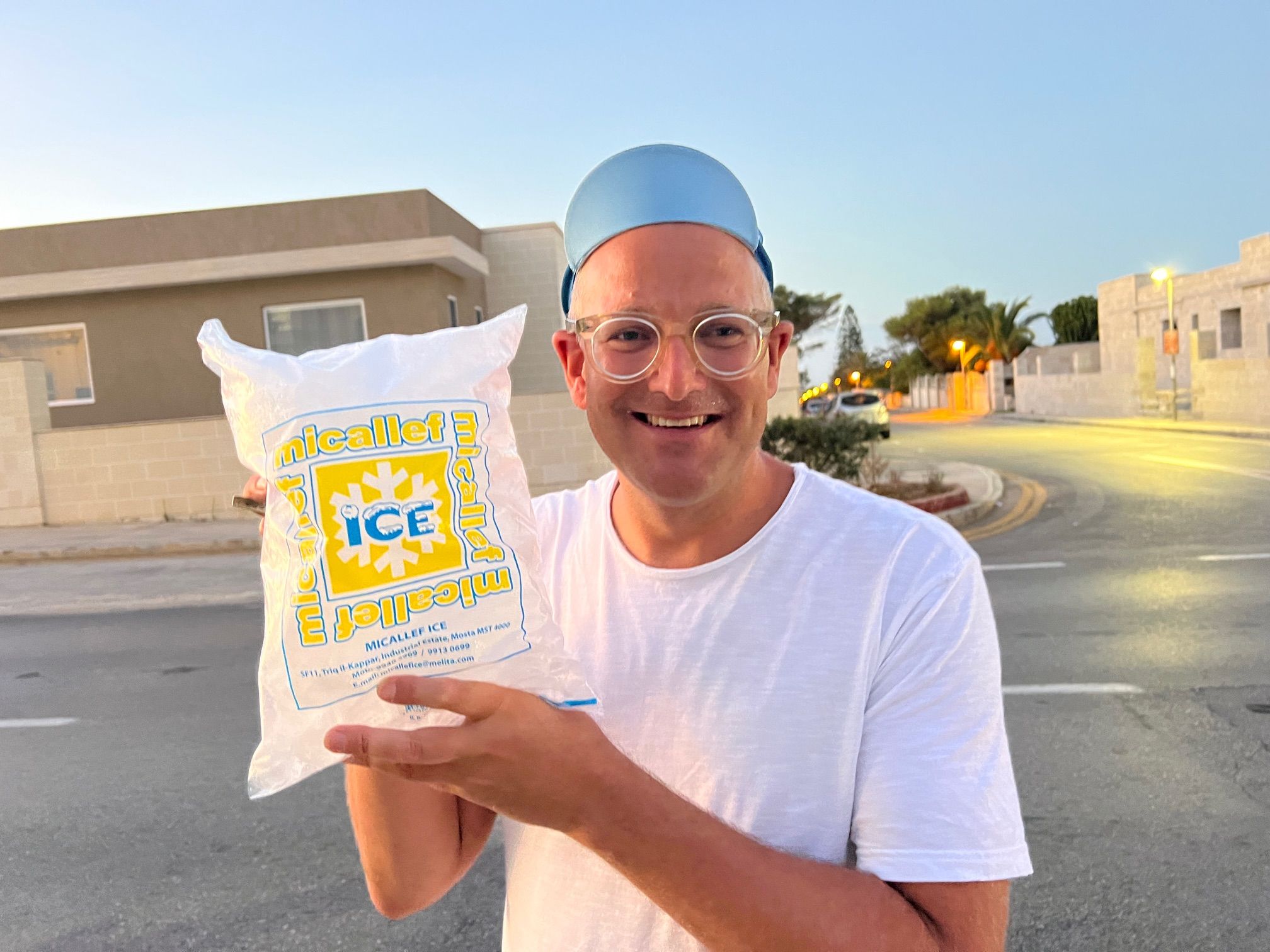 Smiling man in a light blue hat and glasses holds a bag of ice outdoors at dusk.