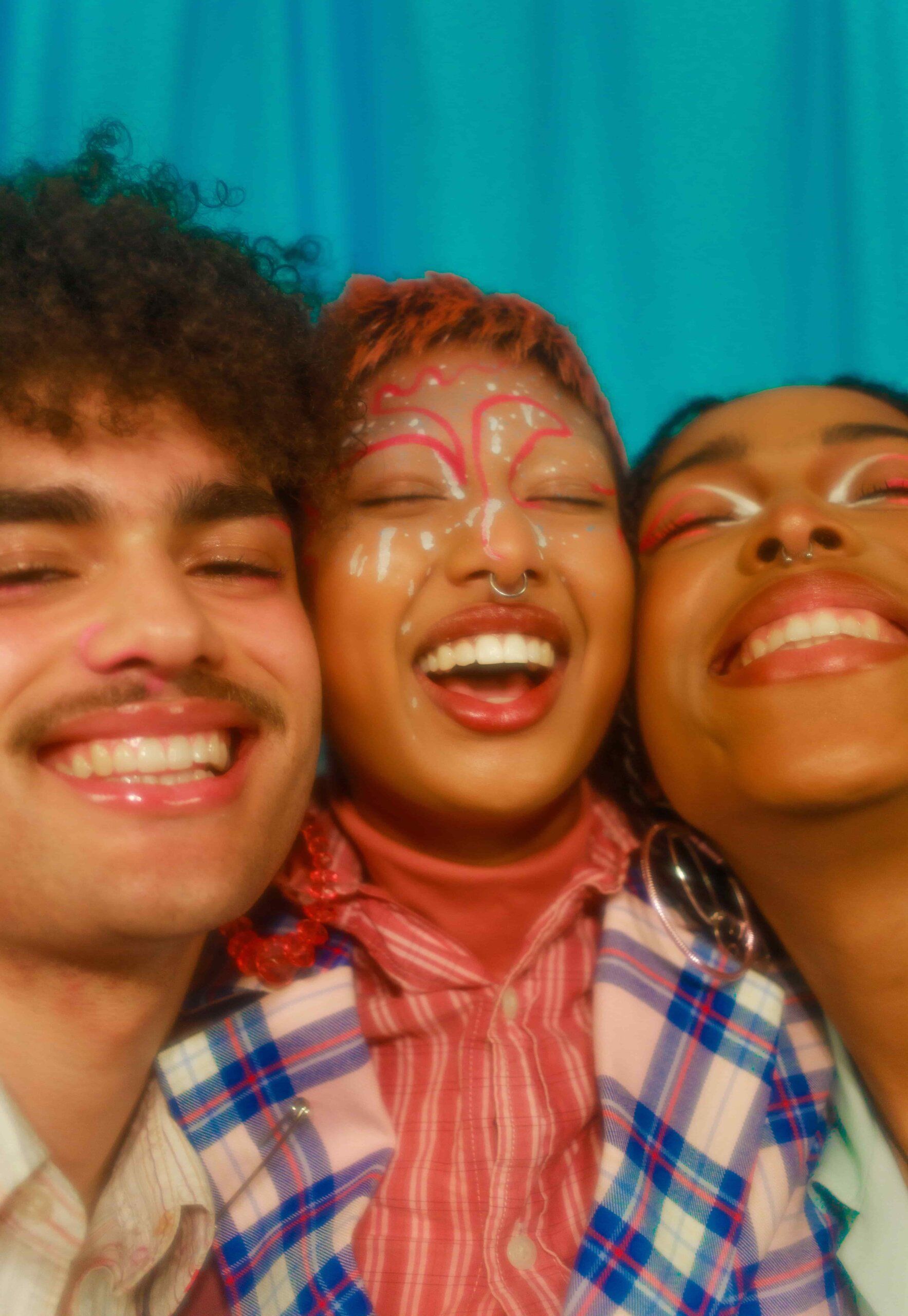 Three diverse young adults with colorful makeup smiling and laughing against a vibrant blue background.