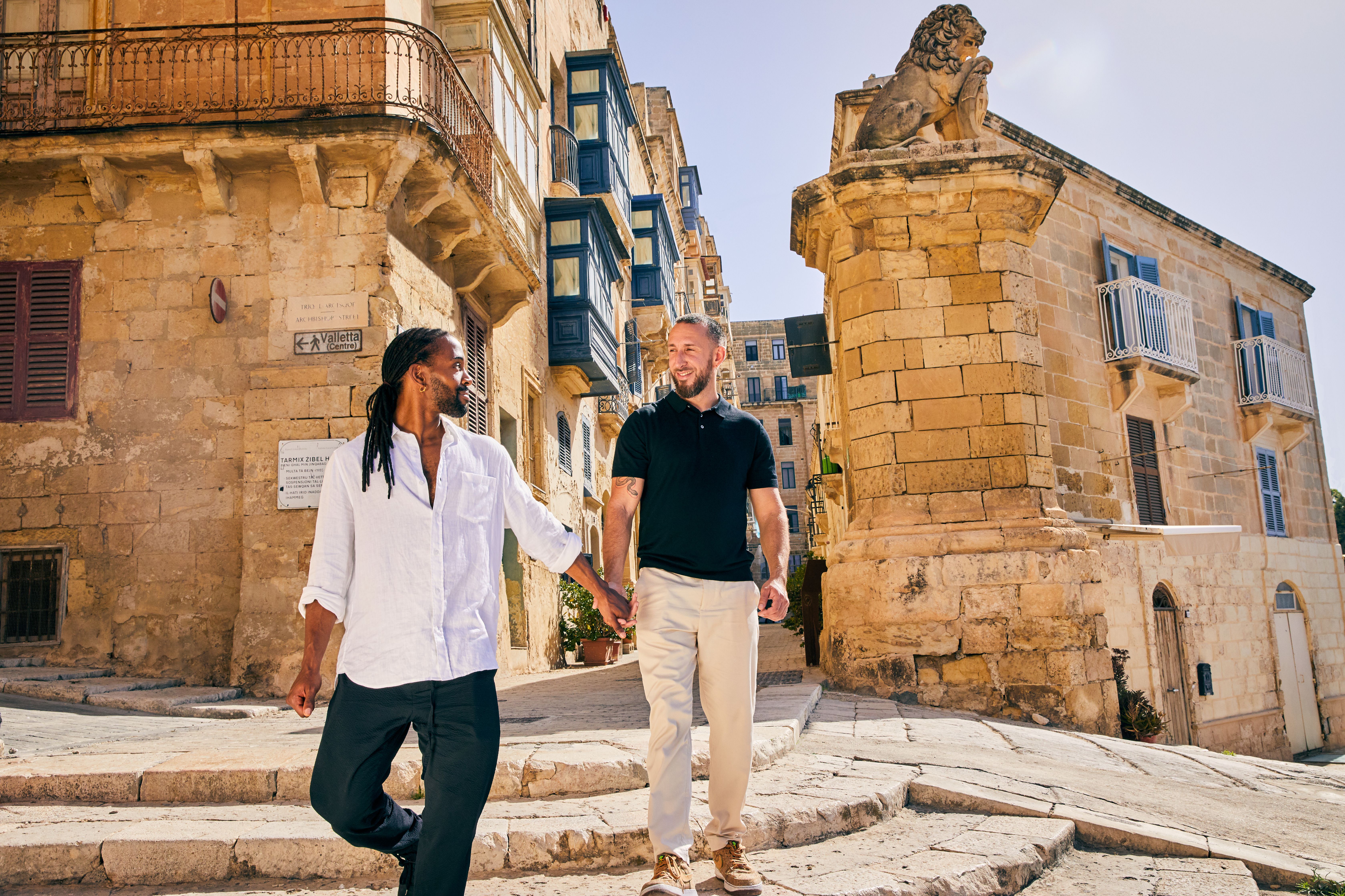 Two men holding hands ascending stone steps in a historic, sunlit Mediterranean city.