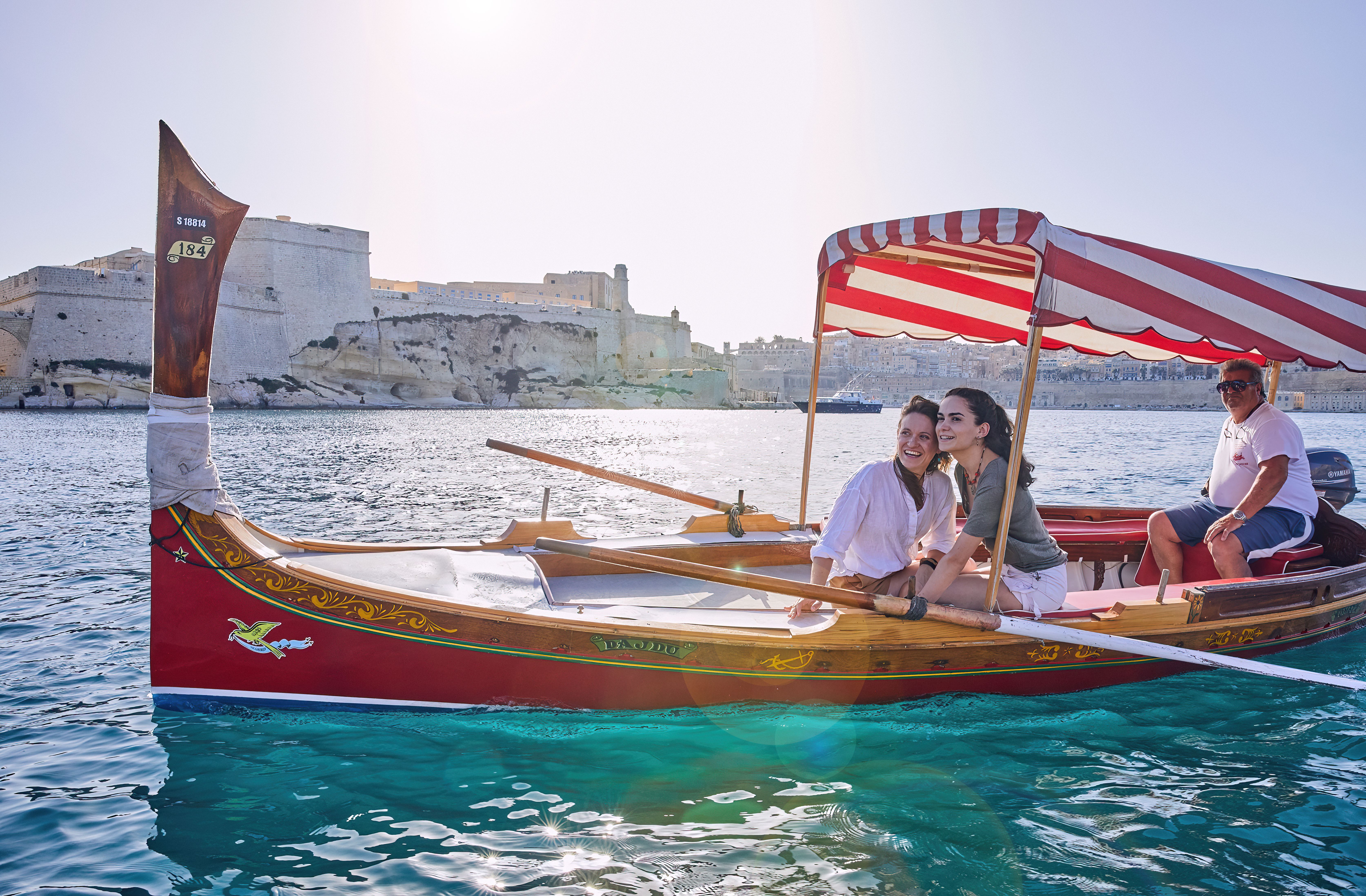 Couple in traditional red boat with striped canopy on blue water, historic fort in background.