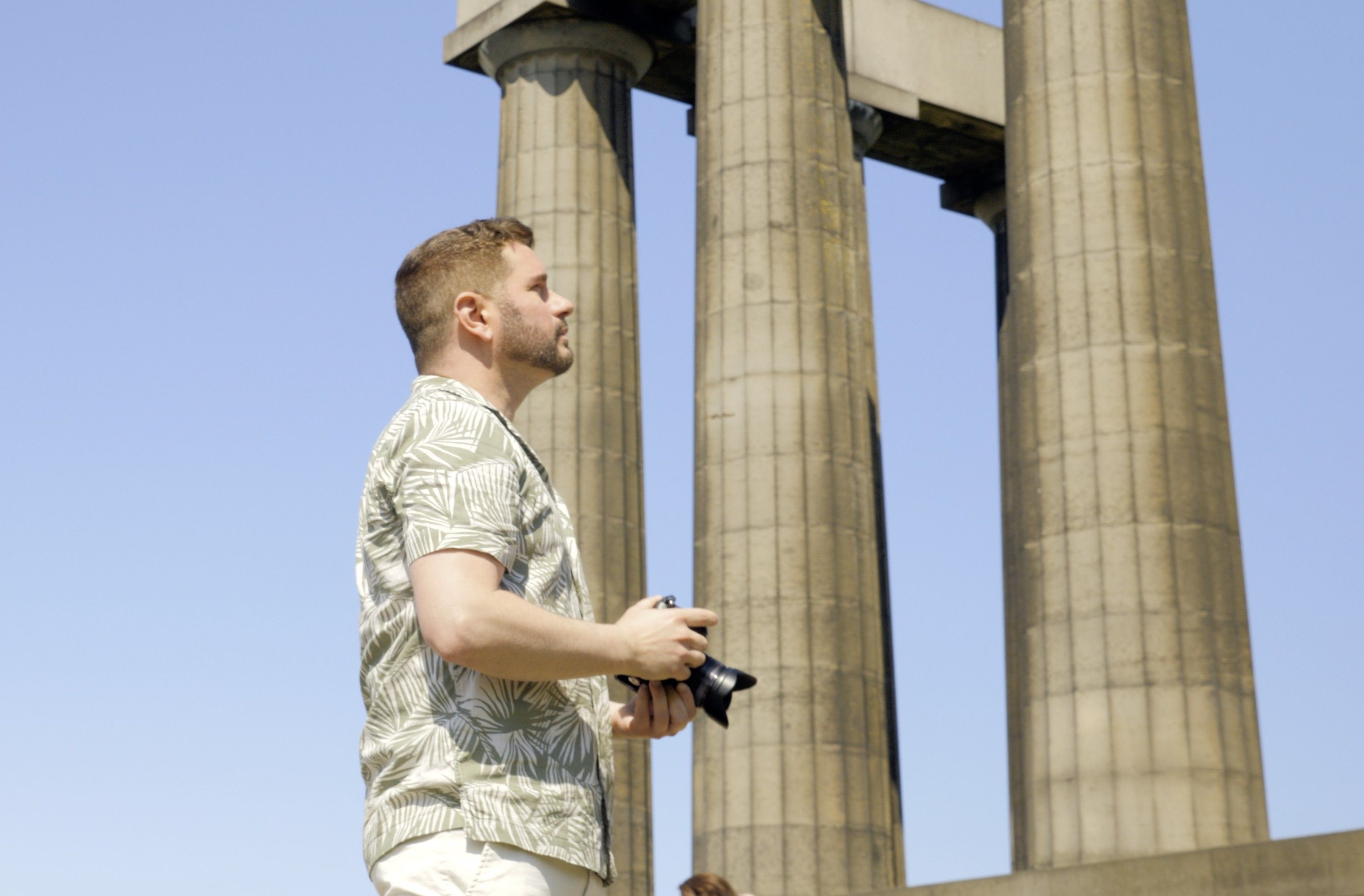 Man with camera looks up at ancient stone columns against a clear blue sky.