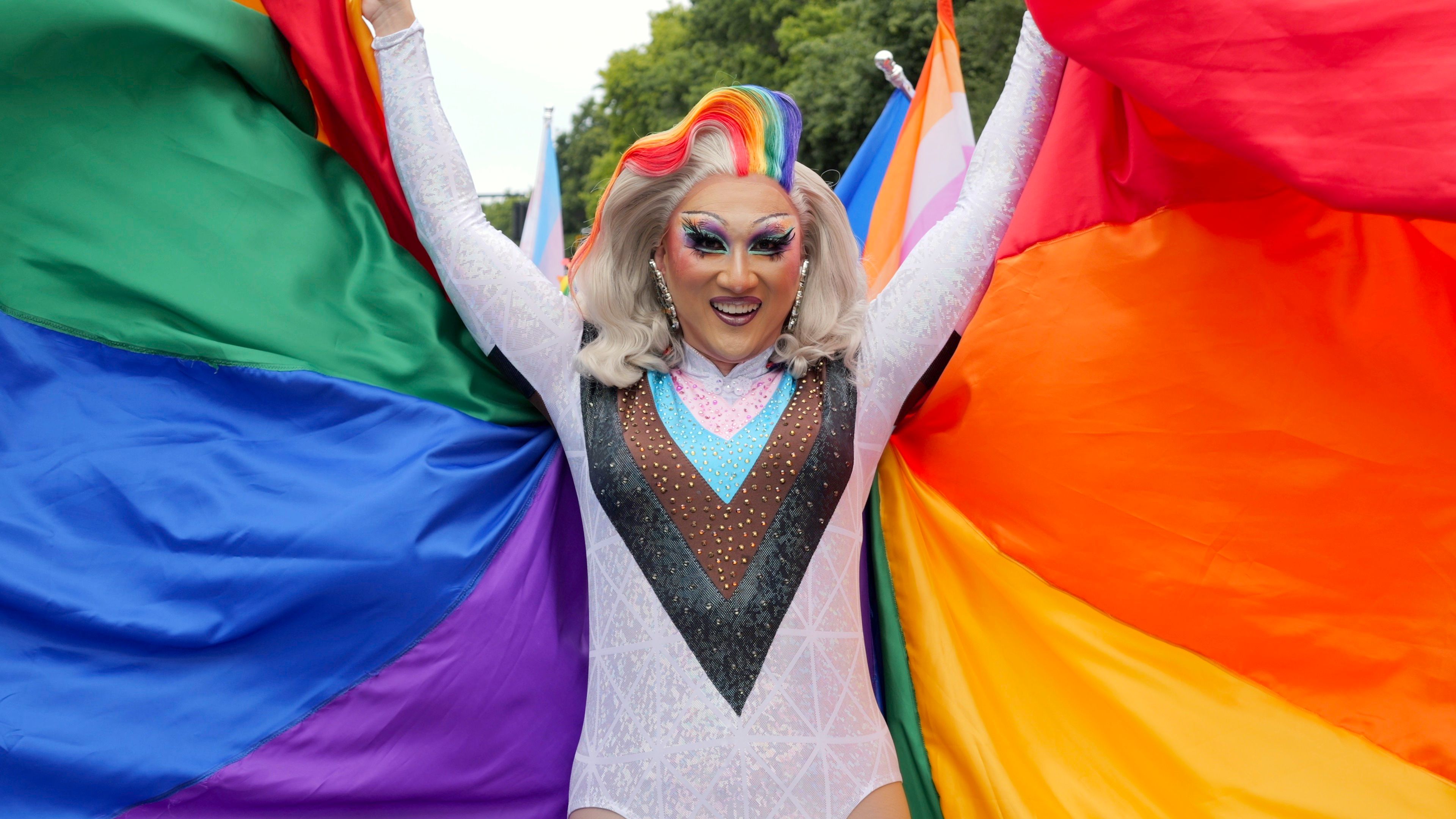 A smiling drag queen holds a large rainbow flag spread wide like colorful wings.