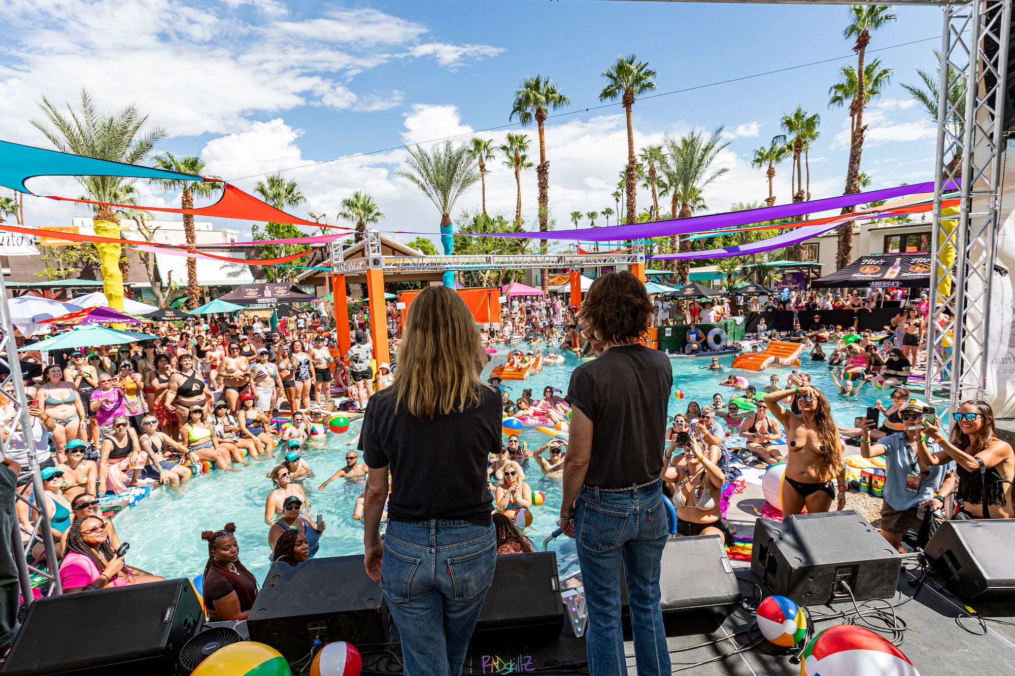 Two people on stage face a large, vibrant outdoor pool party with palm trees.