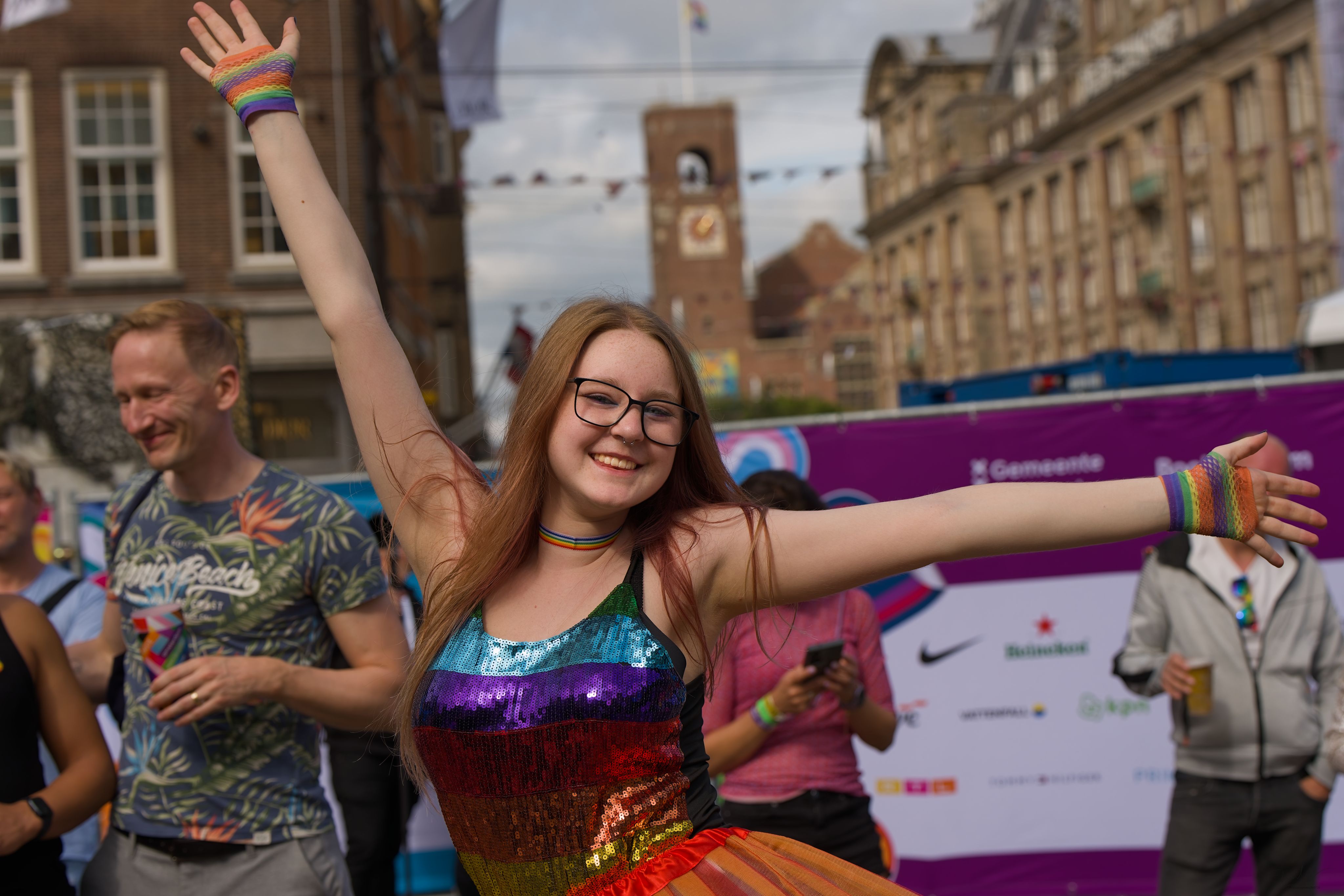 Smiling woman in rainbow top and tutu with raised arms celebrating at an event.