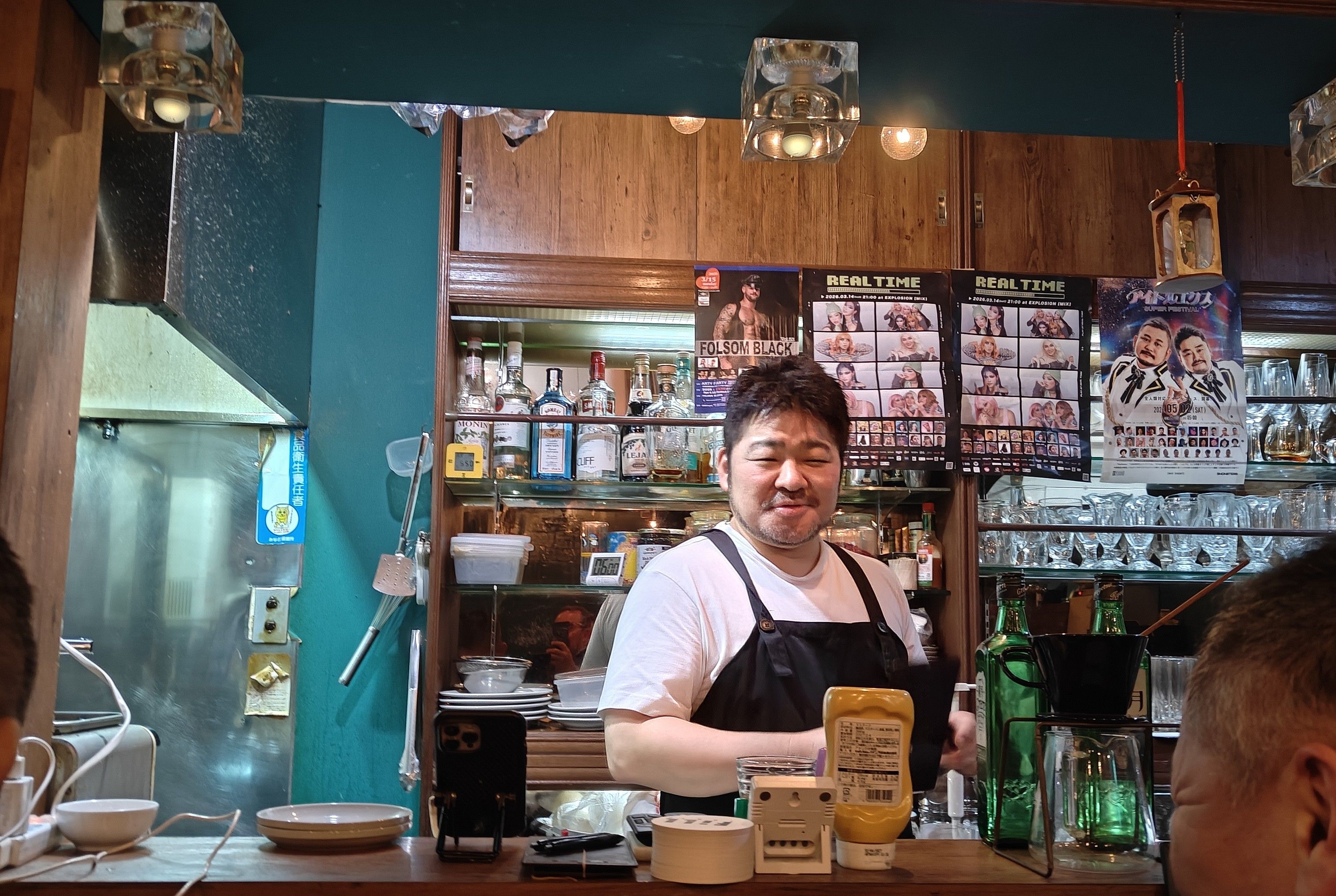 Smiling man in apron behind a bar counter with bottles and posters.