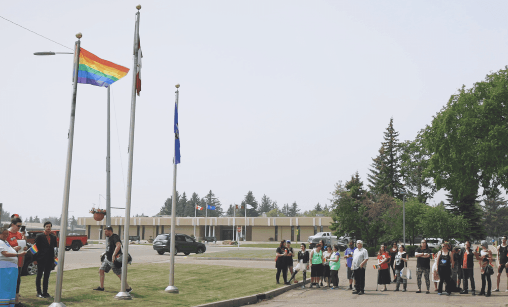 A group of people stand near flagpoles, with a rainbow pride flag waving prominently.