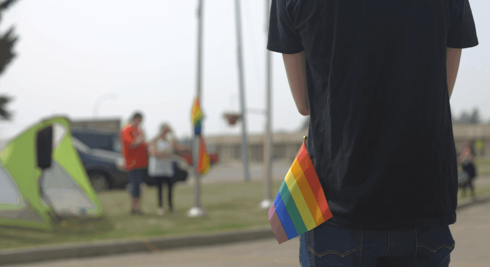 Person from behind with rainbow pride flag in back pocket at outdoor event.