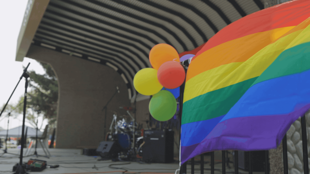 Rainbow flag waves near an outdoor stage with colorful balloons, set for a community event.