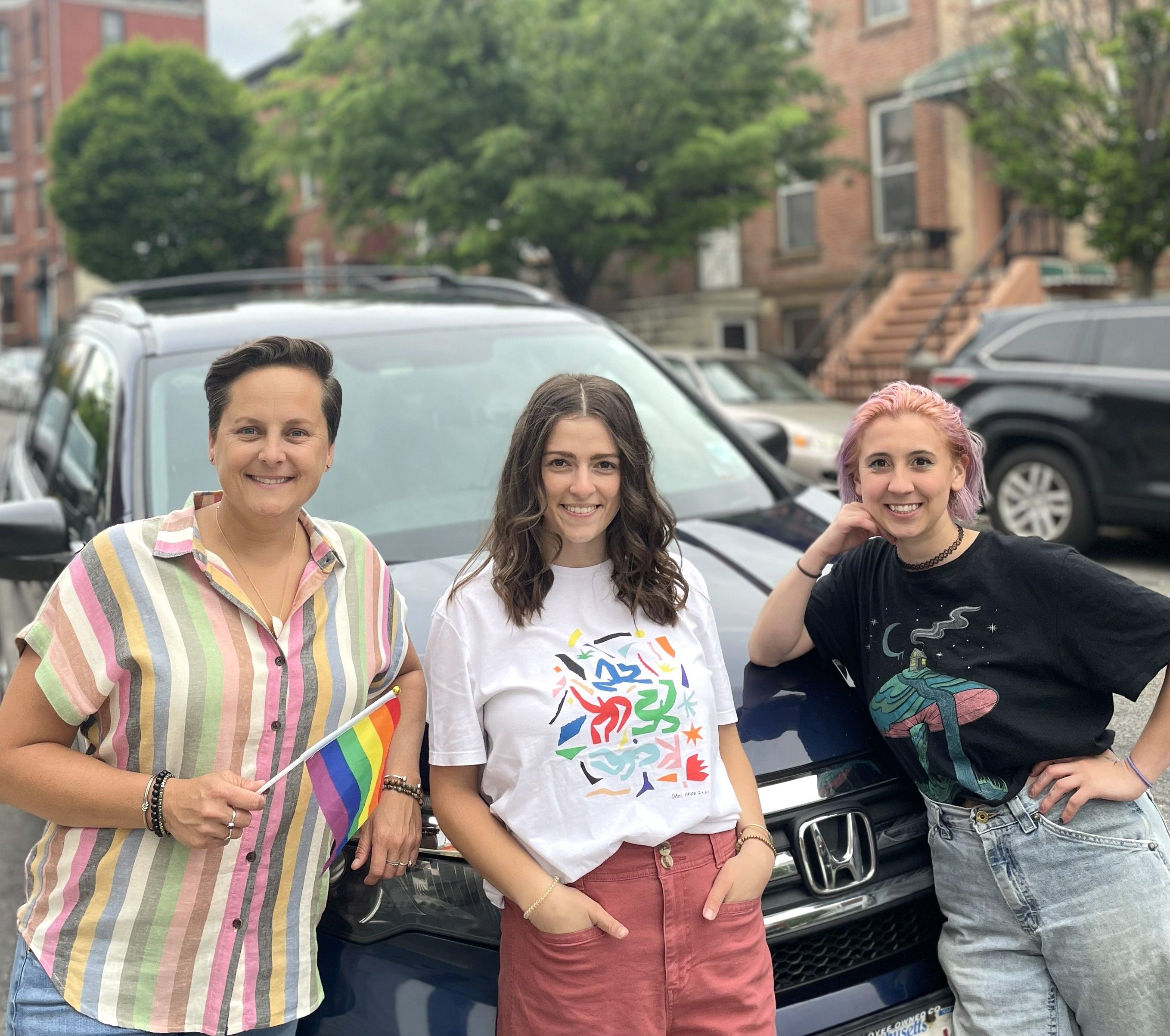 Three women, one holding a rainbow flag, smiling in front of a dark blue SUV.