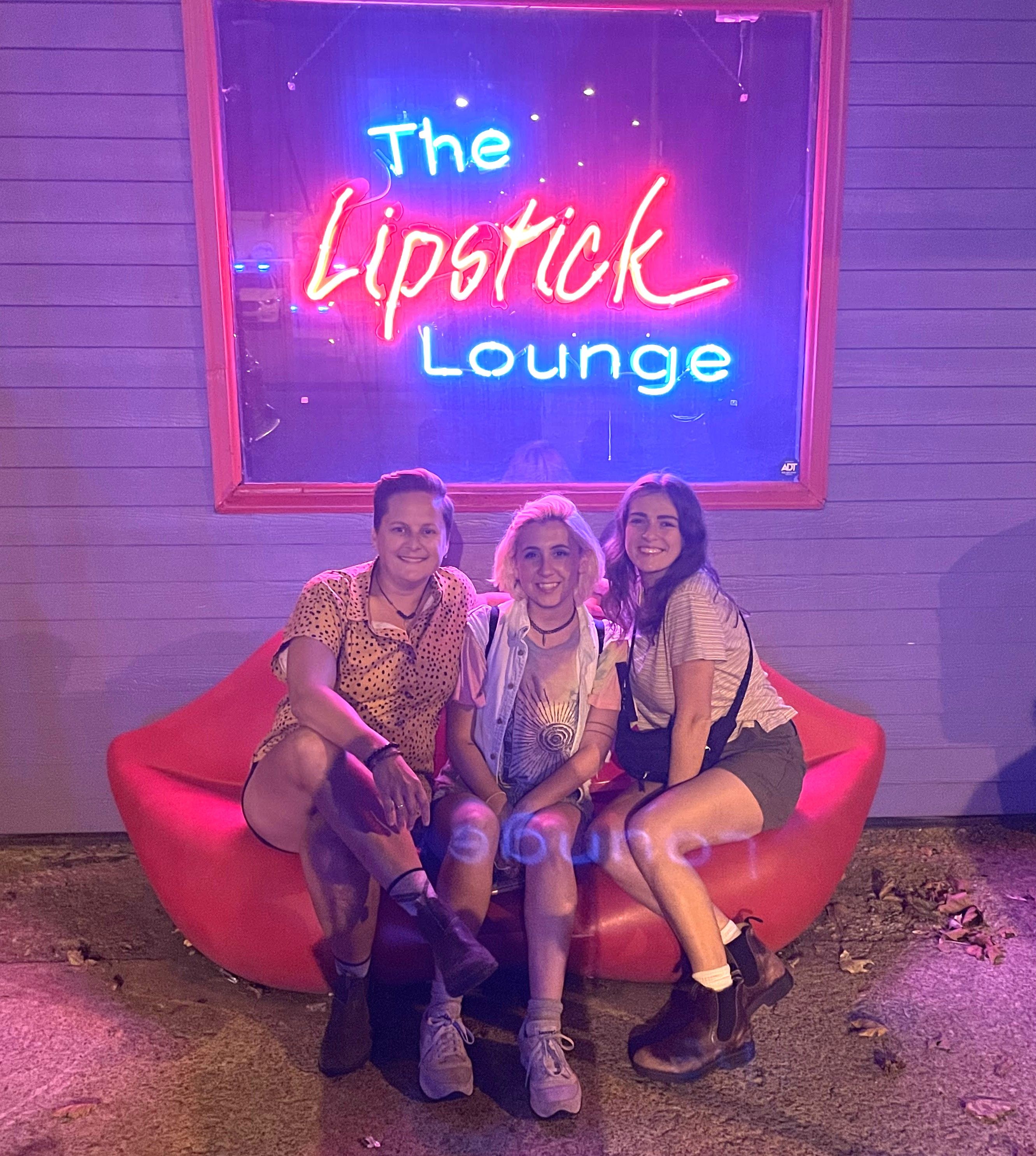 Three friends sit on a red lip couch under a neon 'The Lipstick Lounge' sign.