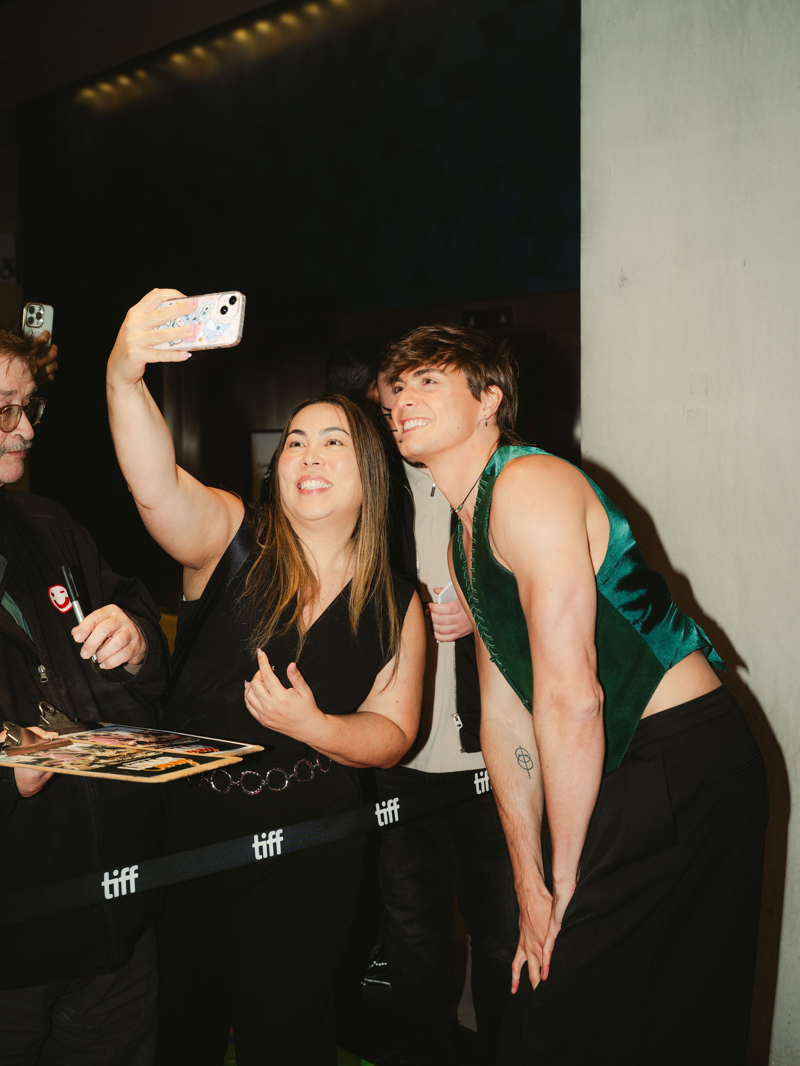 Woman taking a selfie with a man in a green top at a public event.