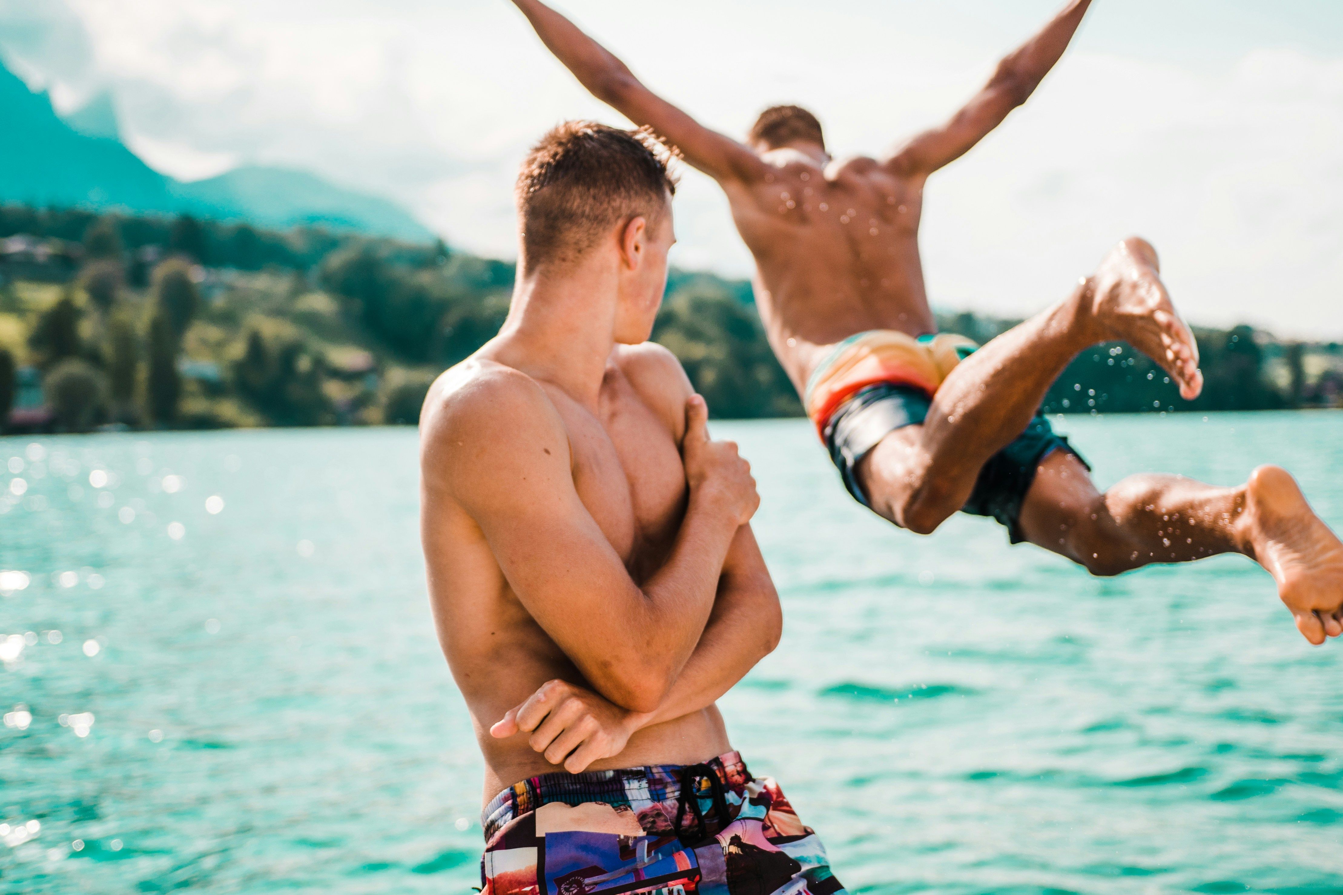 gay travel mistakes Two men at a lake, one jumping into the water as the other watches.