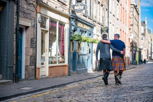 Two men wearing kilts walking arm in arm in a city street