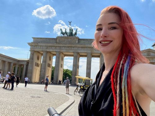 Smiling woman with red hair and dreadlocks taking a selfie in front of Brandenburg Gate, Berlin.
