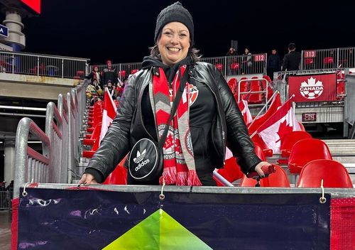Smiling woman in black jacket and red-and-white scarf at a night stadium game.