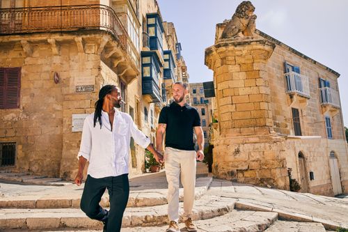 Two men holding hands ascending stone steps in a historic, sunlit Mediterranean city.