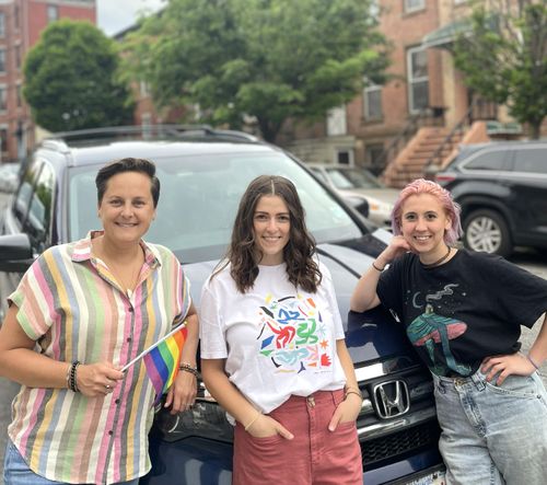 Three women, one holding a rainbow flag, smiling in front of a dark blue SUV.